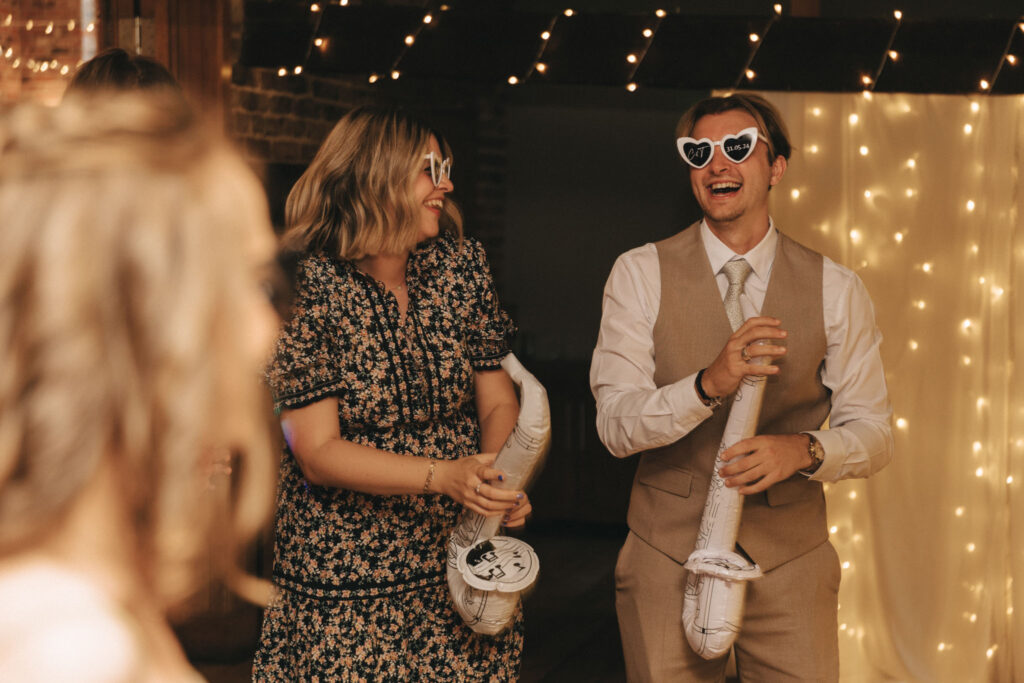 Two people wearing heart-shaped sunglasses enjoy a party. The woman on the left wears a floral dress, and the man on the right is in a suit with a beige vest. Both are holding inflatable guitars and smiling. The background is adorned with twinkling string lights. © Aimee Lince Photography - Wedding photographer in Lincolnshire, Yorkshire & Nottinghamshire
