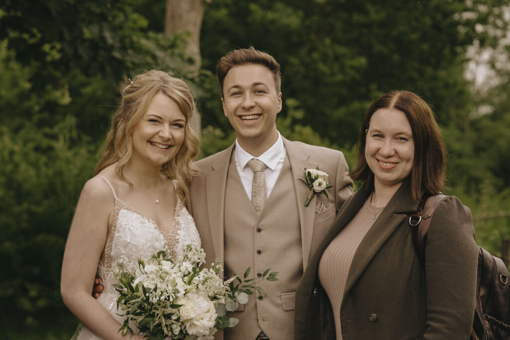 Hall Farm Hotel Wedding Photographer - Courtney & Tom © Aimee Lince Photography Headshot A smiling bride in a lace dress holding white flowers stands next to a groom in a beige suit with a boutonniere, and a woman in a brown coat. They are outdoors with green foliage in the background, all looking happy. © Aimee Lince Photography
