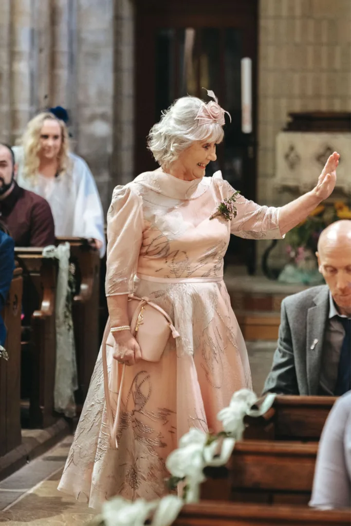 An older woman with white hair waves gracefully as she walks down the flower-adorned church aisle, embodying a mother-daughter moment on your wedding day. In a light pink dress and matching hat, she exudes elegance and charm, ensuring every guest feels the love and joy of this celebration. © Aimee Lince Photography - Wedding photographer in Lincolnshire, Yorkshire & Nottinghamshire