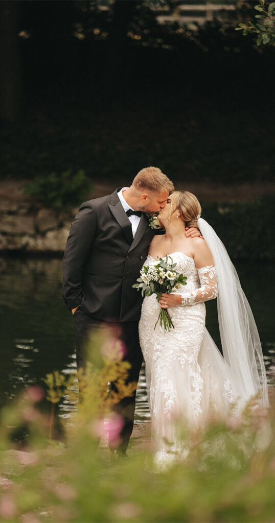 A groom in a black suit and bow tie kisses a bride in a white lace wedding dress, holding a bouquet of white flowers. By the serene lake in Lincolnshire, they are surrounded by greenery and blurred blooms. Her long veil drapes elegantly behind her, completing their perfect moment. © Aimee Lince Photography