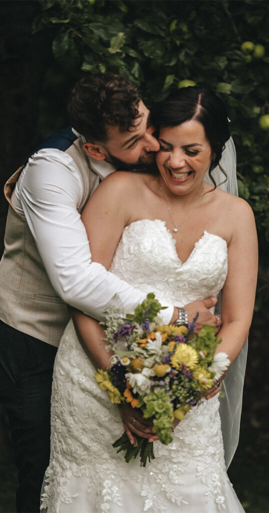 Bride in a white lace wedding dress holds a colorful bouquet while smiling, embraced from behind by the groom wearing a white shirt and beige vest. Both are in front of greenery, looking joyful. © Aimee Lince Photography