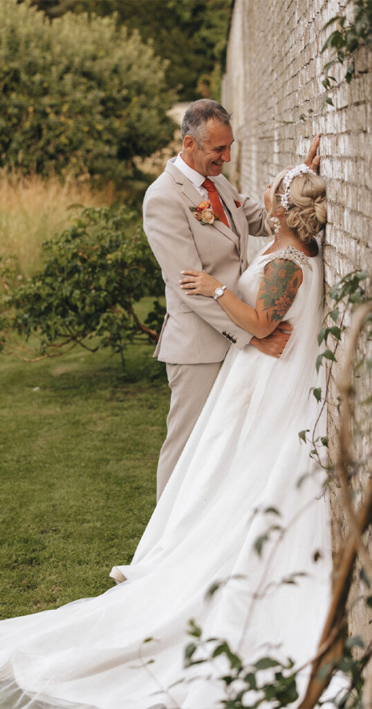 A bride and groom stand intimately by a brick wall. The bride, in a flowing white dress with tattoos, touches the groom’s chest. The groom, in a light beige suit with an orange tie, leans on the wall. Greenery surrounds them, hinting at a garden setting. Both are smiling warmly. © Aimee Lince Photography at Scrivelsby Walled Garden