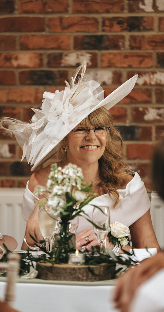 A smiling woman in a white dress and a large white hat with feathers sits at a table decorated with a floral centerpiece. She wears glasses and has long, wavy hair. The background features a brick wall, and there are wine glasses and cutlery on the table. © Aimee Lince Photography