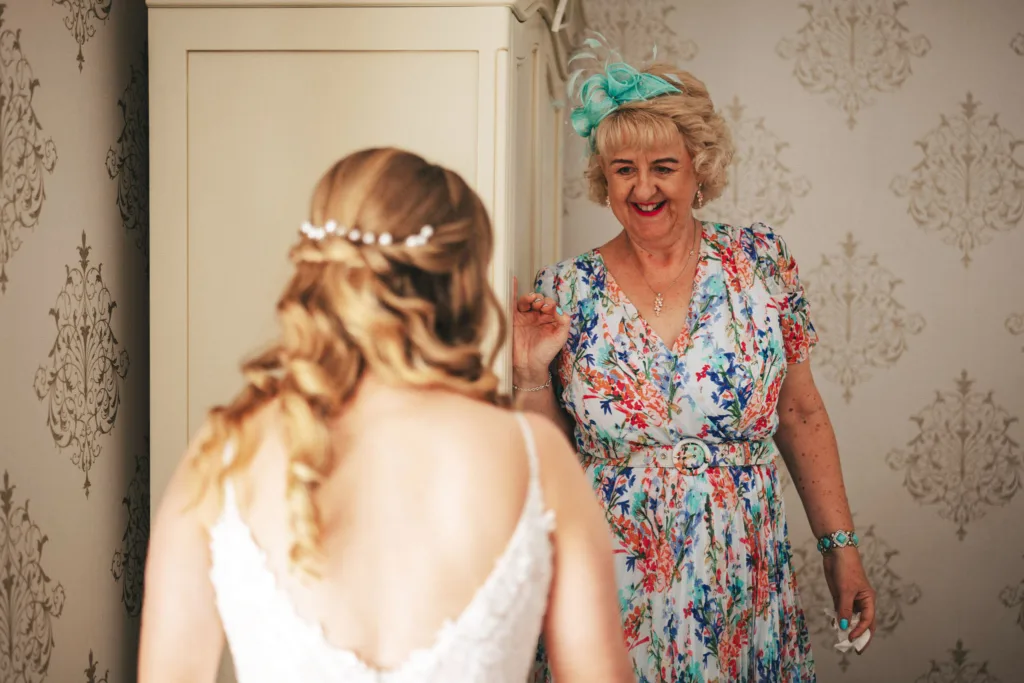 Stallingborough Grange Wedding Photographer - Melissa & Ben © Aimee Lince Photography (83) A bride with long, wavy blonde hair shares a tender mother-daughter moment on her wedding day, facing an older woman in a floral dress and teal hat. The older woman smiles warmly near a cream-colored wardrobe, as the room's vintage wallpaper adds to this cherished scene. © Aimee Lince Photography - Wedding photographer in Lincolnshire, Yorkshire & Nottinghamshire