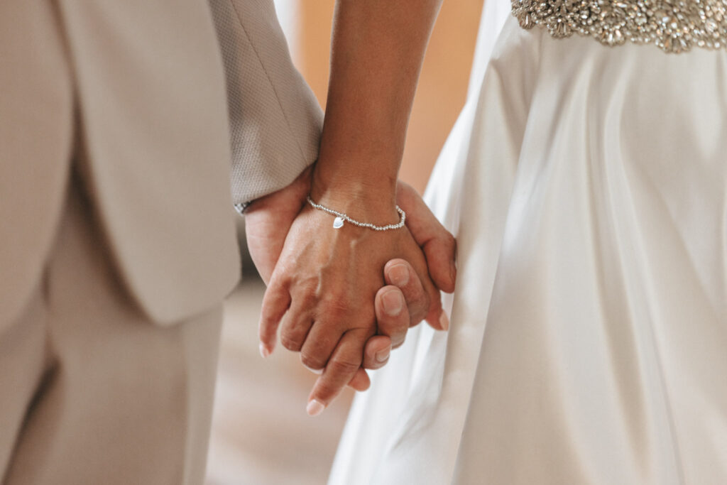 Close-up of a couple holding hands at a wedding. The person on the left is wearing a light gray suit, while the person on the right is in a white dress with intricate beadwork at the waist. A delicate bracelet adorns the right wrist. The background is softly blurred. © Aimee Lince Photography