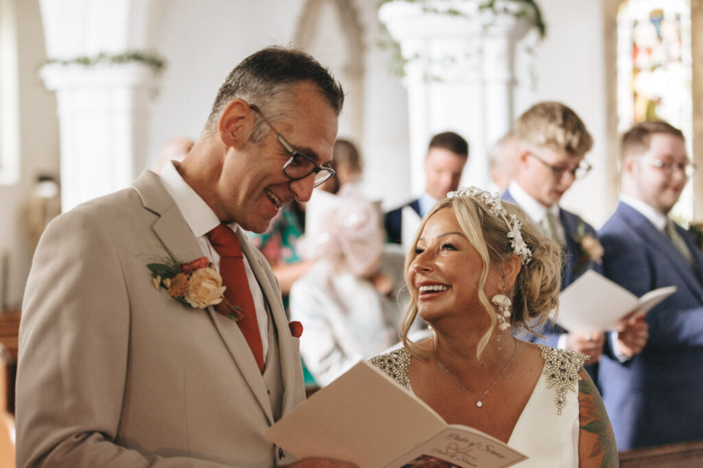 A couple in wedding attire smiles at each other in a church. The groom wears a beige suit with a rose boutonniere and glasses. The bride wears a white gown with beaded shoulders and a floral headpiece. They hold a program. Guests in the background hold similar programs. © Aimee Lince Photography
