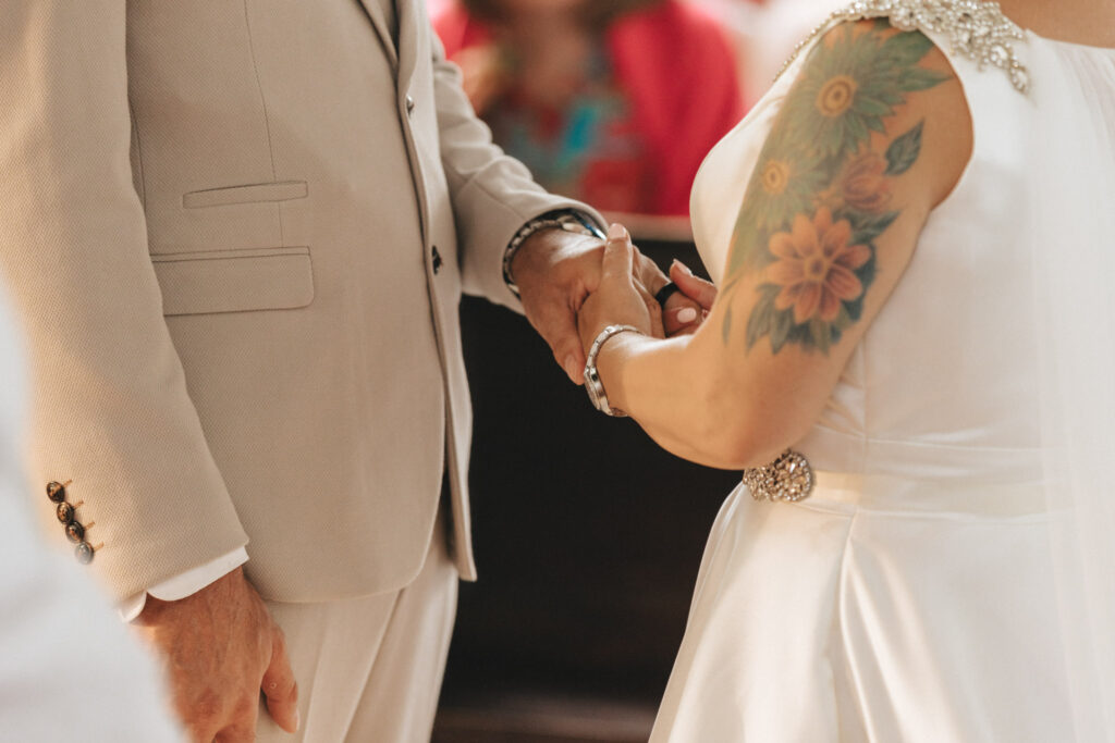 A couple stands facing each other, holding hands. The bride wears a white dress with a sunflower tattoo on her arm and jeweled shoulder details. The groom is in a light beige suit. The background shows blurred, colorful attire of seated guests, suggesting a wedding setting. © Aimee Lince Photography