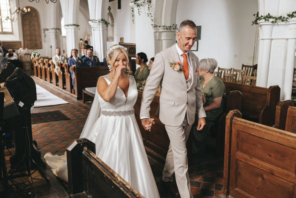 Scrivelsby Walled Garden Wedding Photographer - Simon & Clare © Aimee Lince Photography (191) A bride in a white gown and jeweled headpiece walks down the aisle with an older man in a light gray suit. She is emotional, wiping a tear. Rustic church setting with wooden pews and guests in the background. The aisle has a white runner with floral decorations. © Aimee Lince Photography