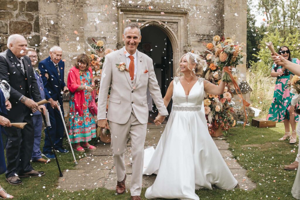 A newlywed couple walks hand in hand, smiling as guests throw confetti. The groom wears a beige suit with an orange tie, while the bride is in a white dress. They are outside a stone building adorned with colorful floral arrangements. People are capturing the moment with cameras and phones. © Aimee Lince Photography