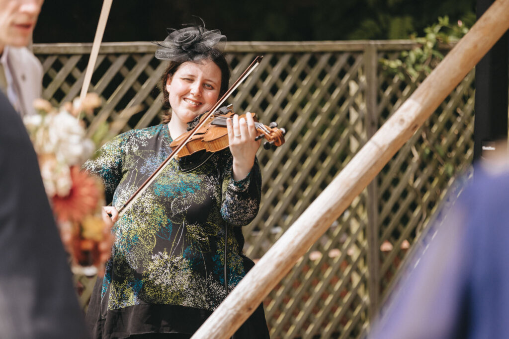 A woman smiles while playing the violin outdoors. She wears a black fascinator and a colorful, patterned top. The wooden structure and lattice fence create a warm, rustic background. Other people are partially visible in the foreground. © Aimee Lince Photography at Scrivelsby Walled Garden
