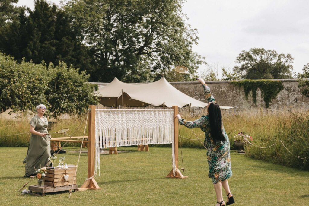 Two women play badminton on a grassy lawn. The net is a decorative macramé hanging. One woman in a floral dress hits the shuttlecock, while the other stands ready. In the background, a cream-colored tent and lush greenery are visible. A wooden crate with flowers is in the foreground. © Aimee Lince Photography at Scrivelsby Walled Garden