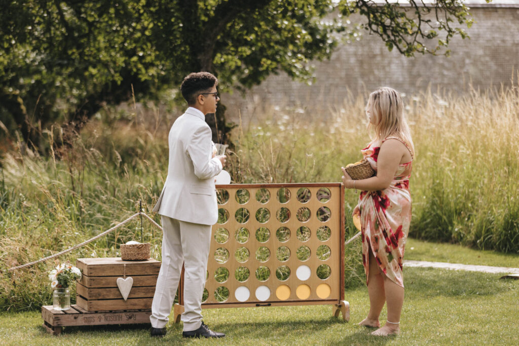 Two people stand outdoors on grass, near a large wooden Connect Four game. One wears a white suit and glasses; the other, a floral dress, barefoot. They're beside wooden crates with a white heart, jars with flowers, near tall grass and a tree. A stone wall is in the background. © Aimee Lince Photography at Scrivelsby Walled Garden