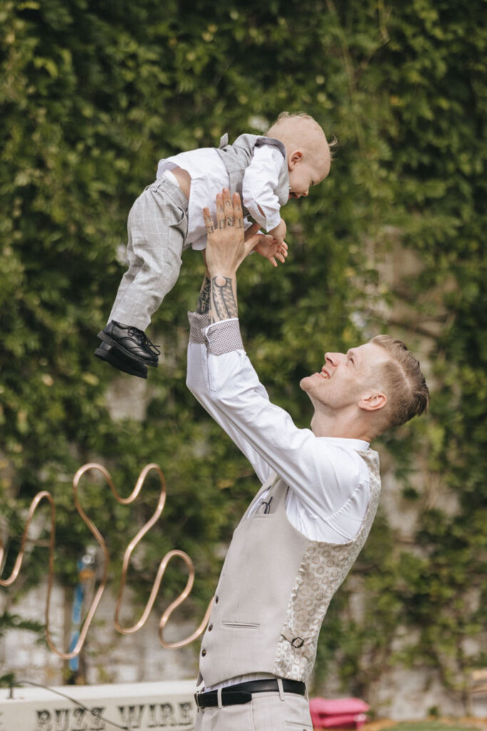 A man in a beige vest and white shirt joyfully lifts a baby in a matching gray outfit. They are outside against a lush green background. The man has visible tattoos and a big smile, while the baby looks down at him cheerfully. A copper sculpture is partially visible behind them. © Aimee Lince Photography at Scrivelsby Walled Garden