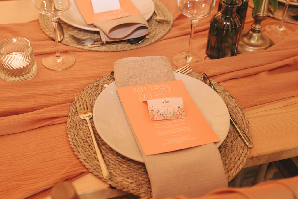 A table setting with woven placemats, white plates, and beige napkins. There's an orange menu card on each plate. Gold cutlery and wine glasses are arranged neatly. An orange fabric runner and small candles in glass holders complete the elegant setup. © Aimee Lince Photography at Scrivelsby Walled Garden