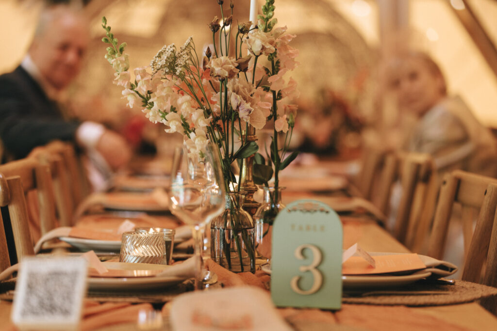 A wooden table is elegantly set for an event, adorned with delicate pink and white flowers in glass vases. Table number 3 is displayed on a teal card. Plates and menus are neatly arranged, with soft ambient lighting creating a warm atmosphere. Two people are seated at the table. © Aimee Lince Photography at Scrivelsby Walled Garden
