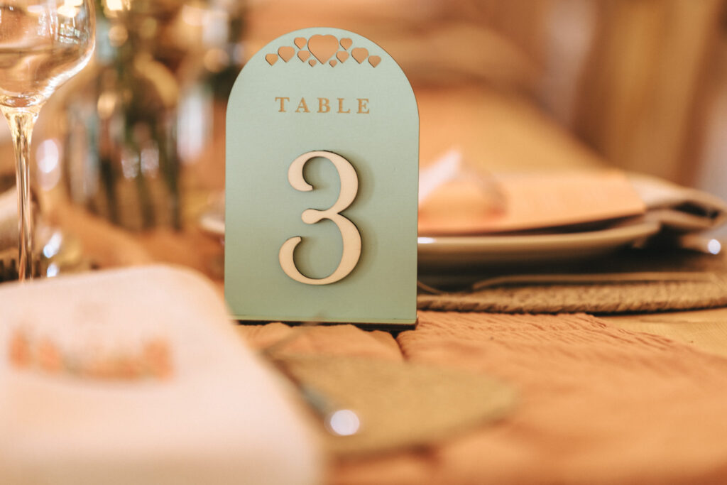 A close-up of a wedding table setting, featuring a table number card with the number 3 in large, white font on a teal background. Decorative heart patterns adorn the top. The table has a soft peach tablecloth, and glasses along with plates are blurred in the background. © Aimee Lince Photography at Scrivelsby Walled Garden