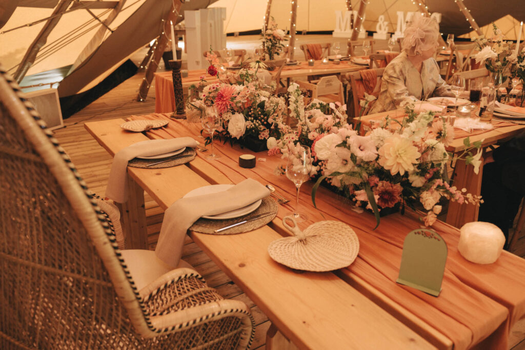 A rustic wedding table setup with wooden tables and wicker chairs. The table is elegantly decorated with floral arrangements of pink and white flowers. Beige napkins are draped over plates. Warm ambient lighting creates a cozy atmosphere, with a backdrop of a tent and wooden flooring. © Aimee Lince Photography at Scrivelsby Walled Garden