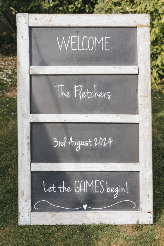 A rustic wooden-framed chalkboard sign stands on grass, surrounded by greenery. The sign reads, "WELCOME The Fletchers 3rd August 2024 let the GAMES begin!" in white handwriting. A small heart is drawn at the bottom. © Aimee Lince Photography at Scrivelsby Walled Garden