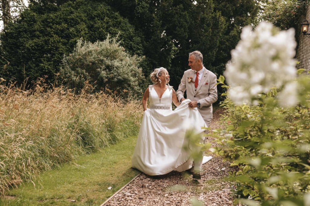 A bride in a white gown and a groom in a beige suit walk joyfully along a garden path surrounded by tall grass and lush greenery. The bride holds her dress, and both are smiling warmly. White flowers are in the foreground, adding to the serene, romantic setting. © Aimee Lince Photography at Scrivelsby Walled Garden