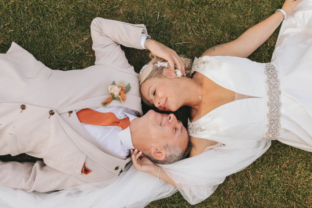 Bride and groom lie on grass, heads touching. The bride wears a white dress with a jeweled belt and veil, her hand gently on the groom's head. The groom wears a beige suit with an orange tie and floral boutonnière. Both have relaxed expressions, gazing into each other's eyes. © Aimee Lince Photography at Scrivelsby Walled Garden