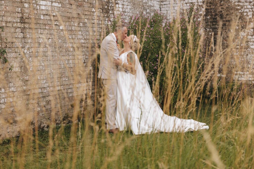A couple in wedding attire share a kiss in front of an old brick wall, partially covered with greenery and flowers. The bride wears a long white dress with a train, and the groom is in a beige suit. Tall grasses in the foreground add a natural frame to the romantic scene. © Aimee Lince Photography at Scrivelsby Walled Garden wedding
