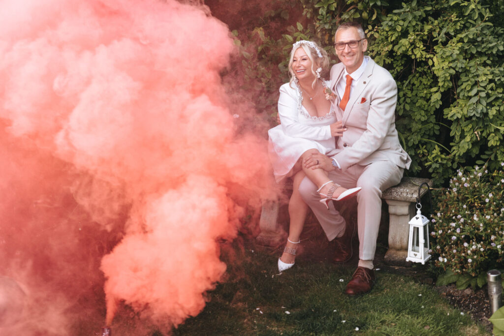 A couple is seated on a stone bench in a garden setting. The woman in a white dress holds the man's hand, who is in a light gray suit with an orange tie. A red smoke bomb to the left fills the scene. Green foliage and a white lantern hang nearby. Both appear joyful and relaxed. © Aimee Lince Photography at Scrivelsby Walled Garden
