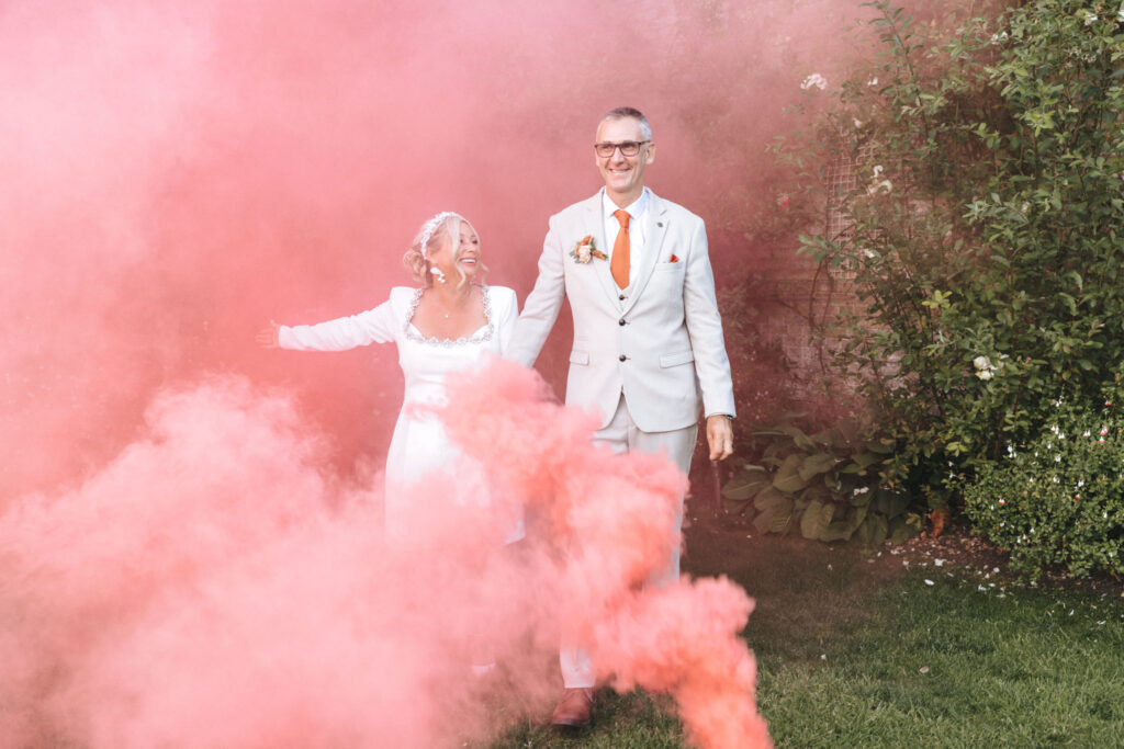A couple in wedding attire walks joyfully through a garden enveloped in pink smoke. The bride wears a long white dress and a tiara, while the groom is in a light gray suit with an orange tie. Both are smiling, and greenery is visible in the background. © Aimee Lince Photography at Scrivelsby Walled Garden