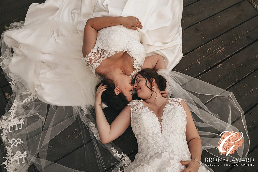 Two brides in lace wedding dresses lie on wooden floorboards, facing each other lovingly. Their veils spread around them as one gently touches the other's face. A "Bronze Award" logo graces the bottom right corner, highlighting this moment's recognition in photography awards. © Aimee Lince Photography - Wedding photographer in Lincolnshire, Yorkshire & Nottinghamshire