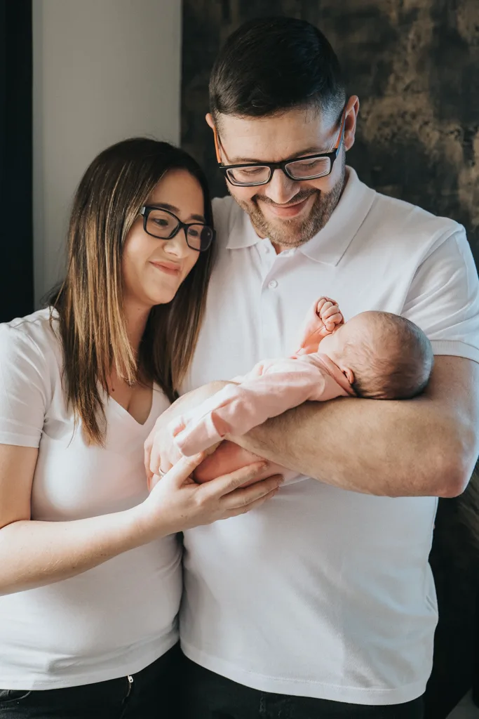 A smiling couple stands closely together holding a newborn. The woman, with long brown hair, and the man, sporting short dark hair and a beard, both wear glasses and white shirts. In this heartwarming photography moment, she gently touches the baby's arm while he cradles their little one in pink. © Aimee Lince Photography - Wedding photographer in Lincolnshire, Yorkshire & Nottinghamshire