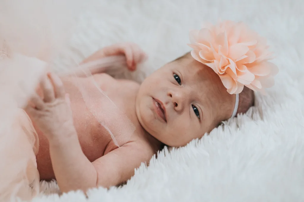 In a captivating display of newborn photography, a baby lies on a soft white blanket, wrapped in sheer pink fabric. She wears a headband with a large peach flower and gazes upward with serenity, her tiny hands gently clutching the fabric—a cherished moment captured perfectly. © Aimee Lince Photography - Wedding photographer in Lincolnshire, Yorkshire & Nottinghamshire