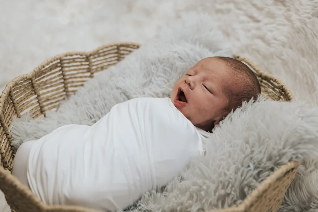 A newborn baby swaddled in a white blanket yawns inside a wicker basket, captured beautifully through photography. Resting on gray, fluffy cushions with a soft, blurred background, this image creates a serene and cozy atmosphere. © Aimee Lince Photography - Wedding photographer in Lincolnshire, Yorkshire & Nottinghamshire