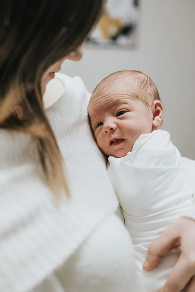 A beautiful piece of newborn photography captures a close-up of a baby wrapped in a soft white blanket, cradled by someone in a white sweater. The baby's eyes are gently closed, and the person's face is tenderly turned toward them. The softly blurred background enhances this tender moment. © Aimee Lince Photography - Wedding photographer in Lincolnshire, Yorkshire & Nottinghamshire