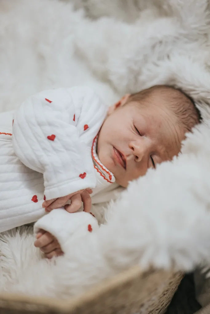 A sleeping newborn is nestled in a fluffy white blanket, captured in exquisite photography. The baby wears a white outfit decorated with small red hearts and red trim around the cuffs and neckline, peacefully lying on their back with eyes closed and hands gently clasped. © Aimee Lince Photography - Wedding photographer in Lincolnshire, Yorkshire & Nottinghamshire