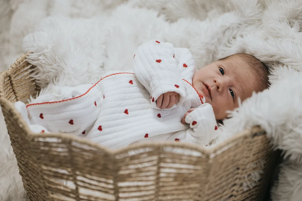 A newborn in a white onesie with red hearts rests in a wicker basket, nestled on a fluffy white blanket. The baby looks upward, hands near their face. This heartwarming photography captures the essence of innocence and comfort. © Aimee Lince Photography - Wedding photographer in Lincolnshire, Yorkshire & Nottinghamshire