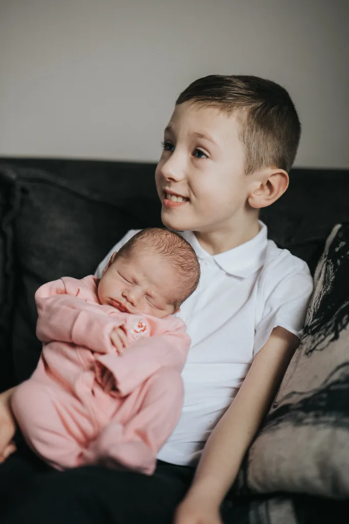 A young boy with short brown hair smiles on a sofa, cradling a sleeping newborn in a pink onesie. His white shirt contrasts softly against the neutral-toned wall and cushion behind. This tender moment captures the essence of family photography. © Aimee Lince Photography - Wedding photographer in Lincolnshire, Yorkshire & Nottinghamshire