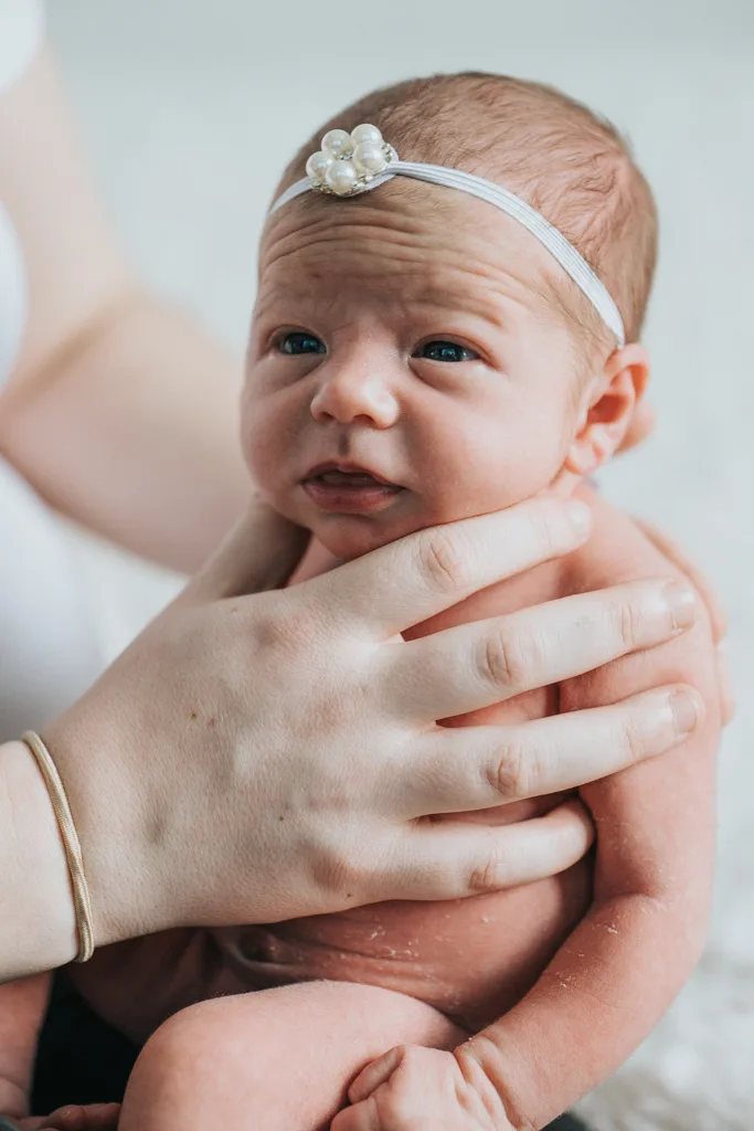 A newborn, captured in serene photography, wears a white headband with a pearl decoration and looks upward. A woman's hand, adorned with a thin bracelet, gently supports the baby. The softly blurred background highlights the baby's delicate features and peach-colored skin. © Aimee Lince Photography - Wedding photographer in Lincolnshire, Yorkshire & Nottinghamshire