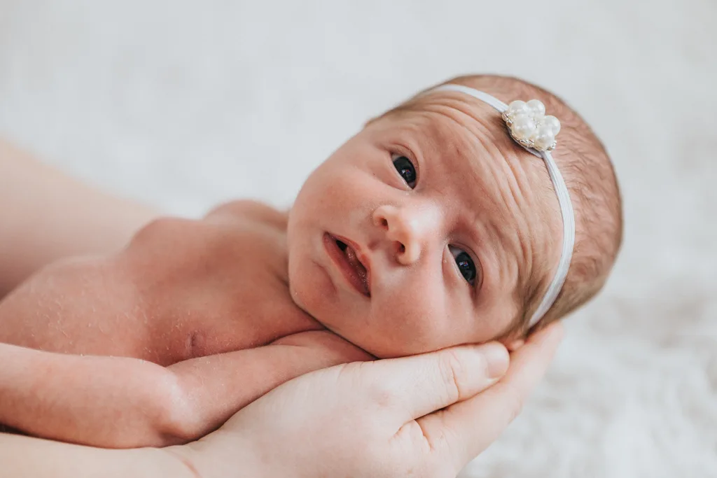 Close-up of a newborn baby lying on a soft, white blanket. The baby, wearing a delicate headband with a pearl flower, has eyes open and is being gently supported by adult hands under its head. This exquisite photography captures the tenderness and care of Scunthorpe's newest resident. © Aimee Lince Photography - Wedding photographer in Lincolnshire, Yorkshire & Nottinghamshire