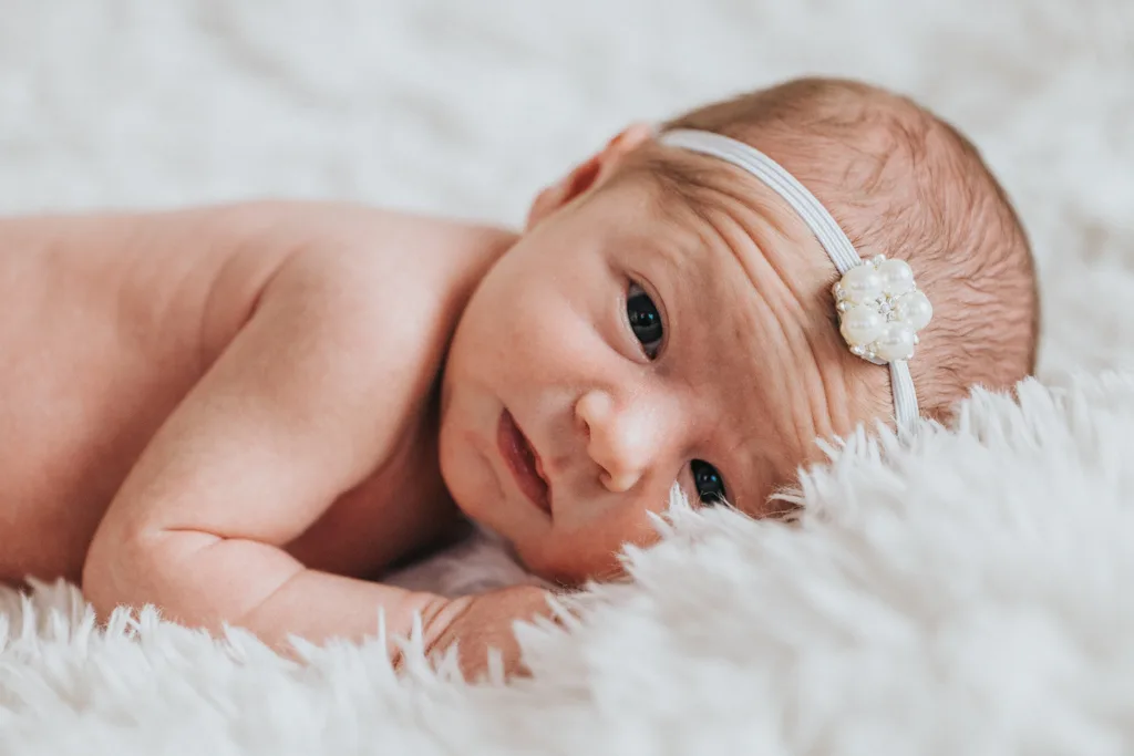 A newborn baby lies on a fluffy white blanket, captured in exquisite photography. Wearing a white headband adorned with a small flower, the infant's light skin and open eyes gaze serenely at the camera. The calm setting embodies a soft-focused elegance. © Aimee Lince Photography - Wedding photographer in Lincolnshire, Yorkshire & Nottinghamshire
