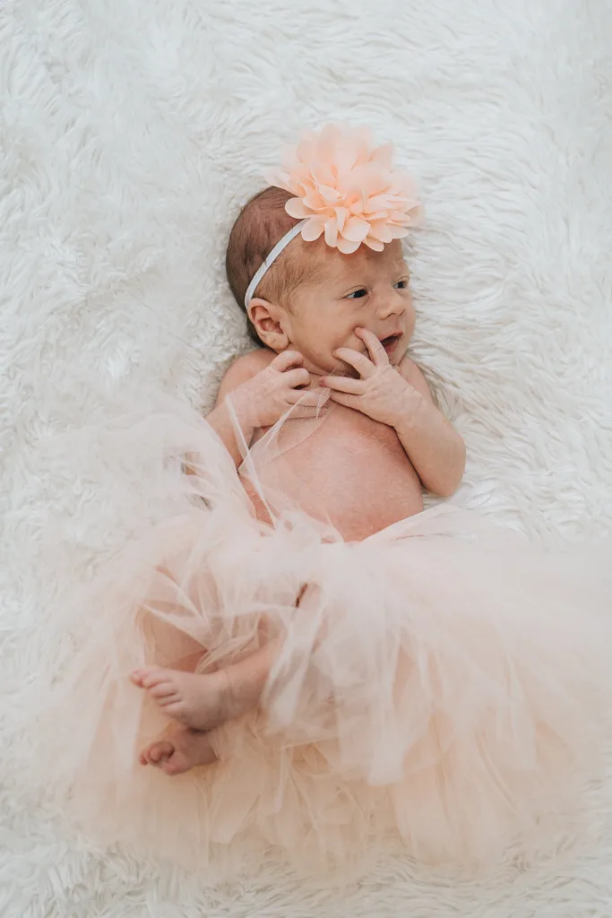 A newborn baby lies on a soft white blanket, capturing the serene beauty that Scunthorpe photography is known for. The baby wears a peach tulle skirt and matching headband adorned with a large peach flower, exuding calmness as their tiny feet peek out playfully. © Aimee Lince Photography - Wedding photographer in Lincolnshire, Yorkshire & Nottinghamshire