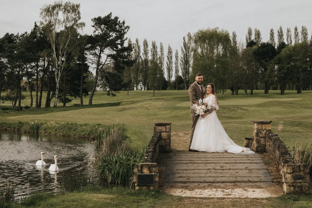 A bride and groom stand on a small stone bridge in a lush park setting, surrounded by family. The bride holds a bouquet and wears a white dress, while the groom wears a brown suit. Behind them are a pond with two swans and trees against grassy fields under a cloudy sky, creating memories worthy of any journal. © Aimee Lince Photography - Wedding photographer in Lincolnshire, Yorkshire & Nottinghamshire