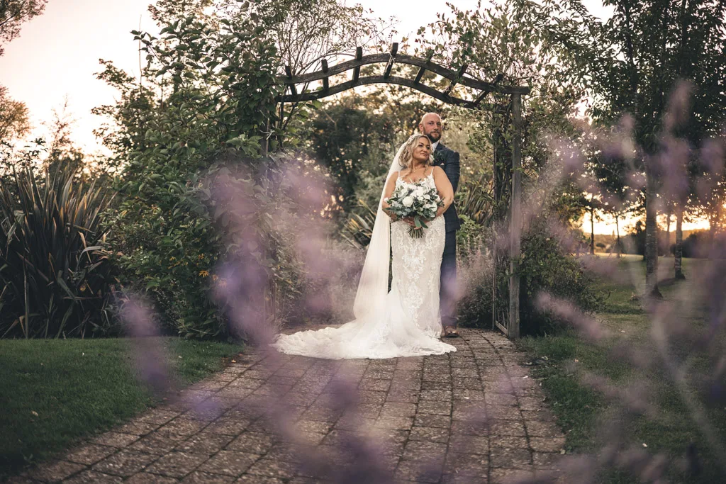 A bride and groom, united in their engagement, stand together under a wooden archway in a garden at sunset. The bride, in a lace gown and veil, holds a bouquet of flowers. The groom is in a dark suit. Purple flowers blur the foreground, while trees and greenery surround them, creating a romantic atmosphere. © Aimee Lince Photography - Wedding photographer in Lincolnshire, Yorkshire & Nottinghamshire