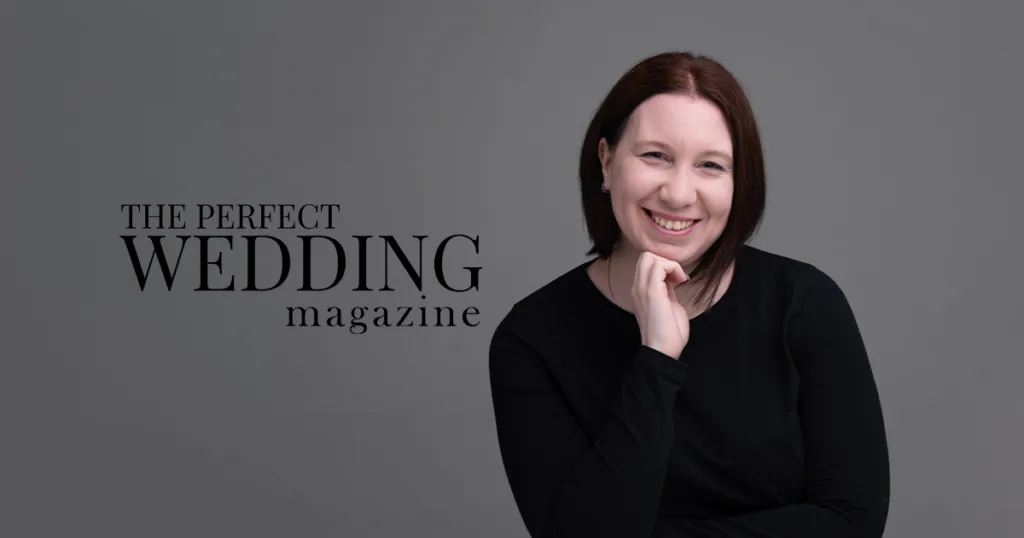 A writer with shoulder-length dark brown hair smiles while resting their chin on their hand, embodying the charm of "THE PERFECT WEDDING" magazine. Against a gray backdrop, they wear a black top. Exciting news awaits to the left in bold black letters. © Aimee Lince Photography - Wedding photographer in Lincolnshire, Yorkshire & Nottinghamshire