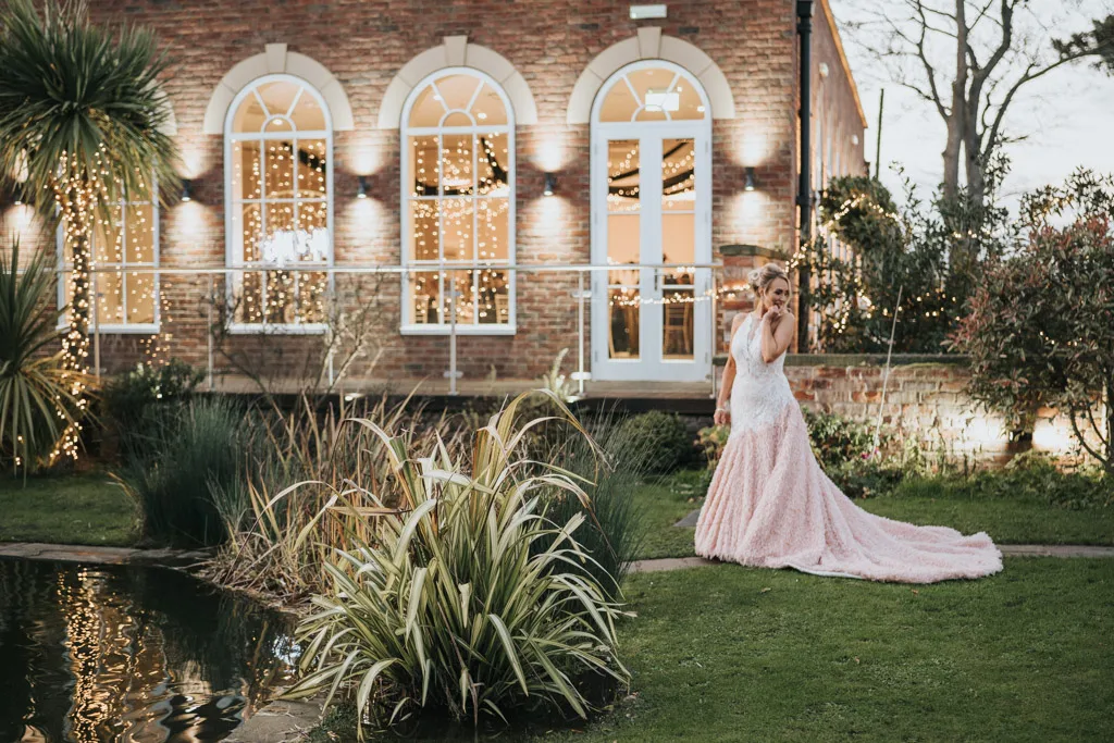 Bride in a sleeveless pink ball gown poses elegantly beside a pond as the photographer captures the moment. The dress features a long train. A brick building with arched, light-adorned windows and lush greenery enhances the serene ambiance, making it a cherished memory for her family. © Aimee Lince Photography - Wedding photographer in Lincolnshire, Yorkshire & Nottinghamshire
