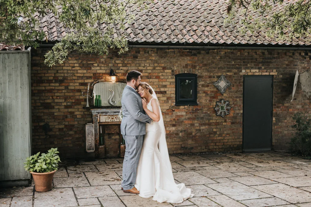 A photographer captures a couple embracing in front of a rustic brick wall. The groom, in a light gray suit, gently holds the bride in her white gown and veil. Standing on a stone patio with potted plants nearby, they add charm to the portfolio's scene with mirrors and metal art adorning the wall. © Aimee Lince Photography - Wedding photographer in Lincolnshire, Yorkshire & Nottinghamshire