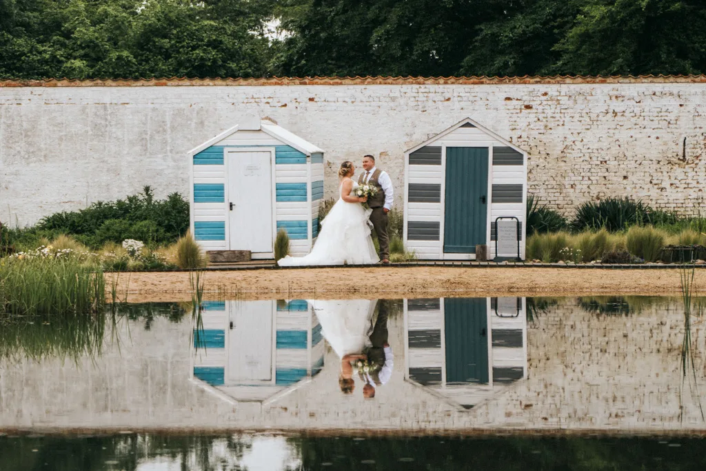 A bride and groom stand in front of two striped beach huts—one light blue and white, the other dark blue and gray—against a white brick wall. Captured by the photographer's keen eye, their image is reflected in a calm pond surrounded by greenery and reeds, perfect for any portfolio. © Aimee Lince Photography - Wedding photographer in Lincolnshire, Yorkshire & Nottinghamshire