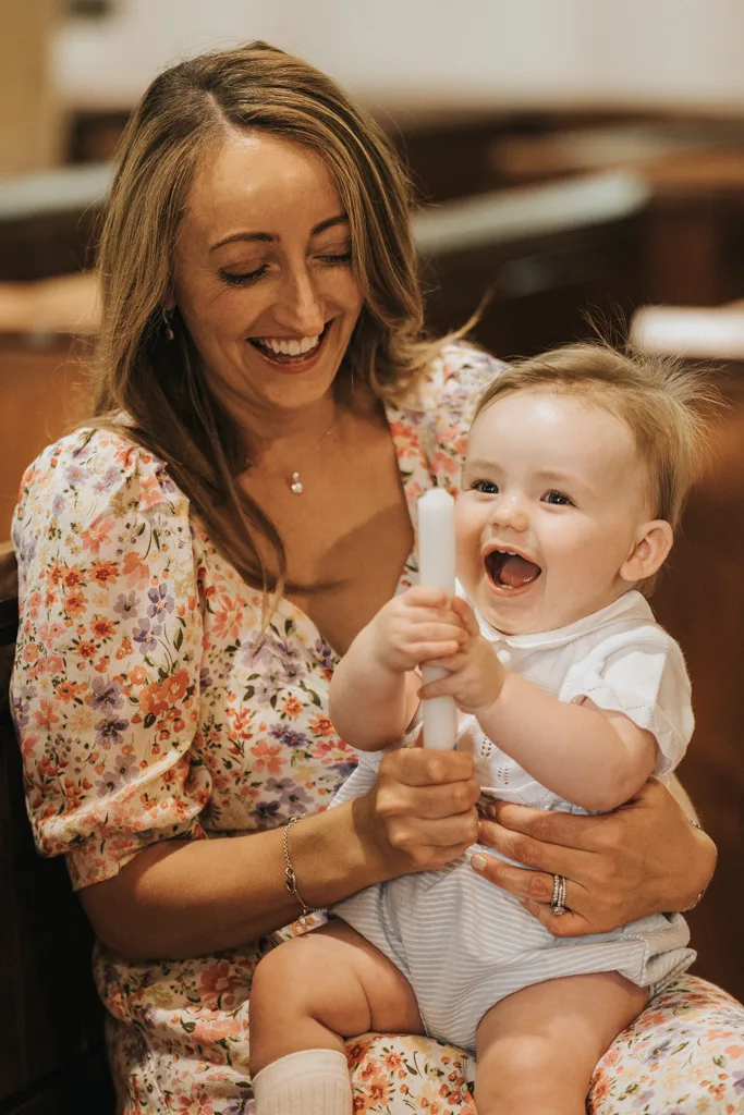 A smiling woman in a floral dress holds a happy baby dressed in white, celebrating their christening. The baby, clutching a white candle and laughing, sits indoors with her. The atmosphere is joyful and warm, capturing the spirit of this special day. © Aimee Lince Photography - Wedding photographer in Lincolnshire, Yorkshire & Nottinghamshire