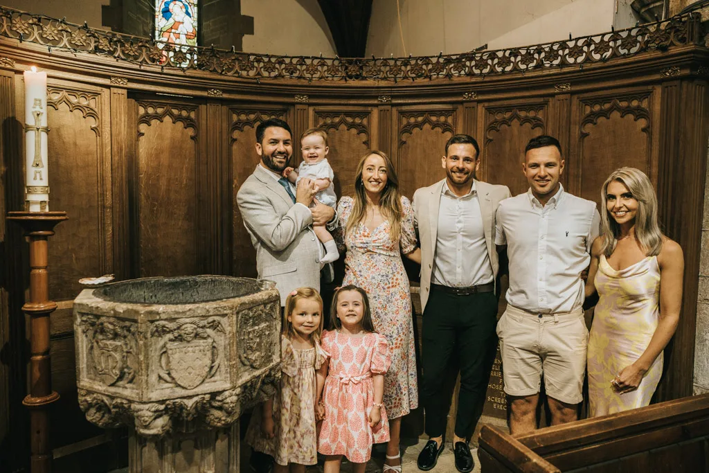 A group of seven people, including three men and three women, gather around a stone baptismal font inside a church with wooden paneling in Grimsby. Two of the women hold children during the christening. A lit candle stands nearby, while a stained glass window is partially visible above. © Aimee Lince Photography - Wedding photographer in Lincolnshire, Yorkshire & Nottinghamshire