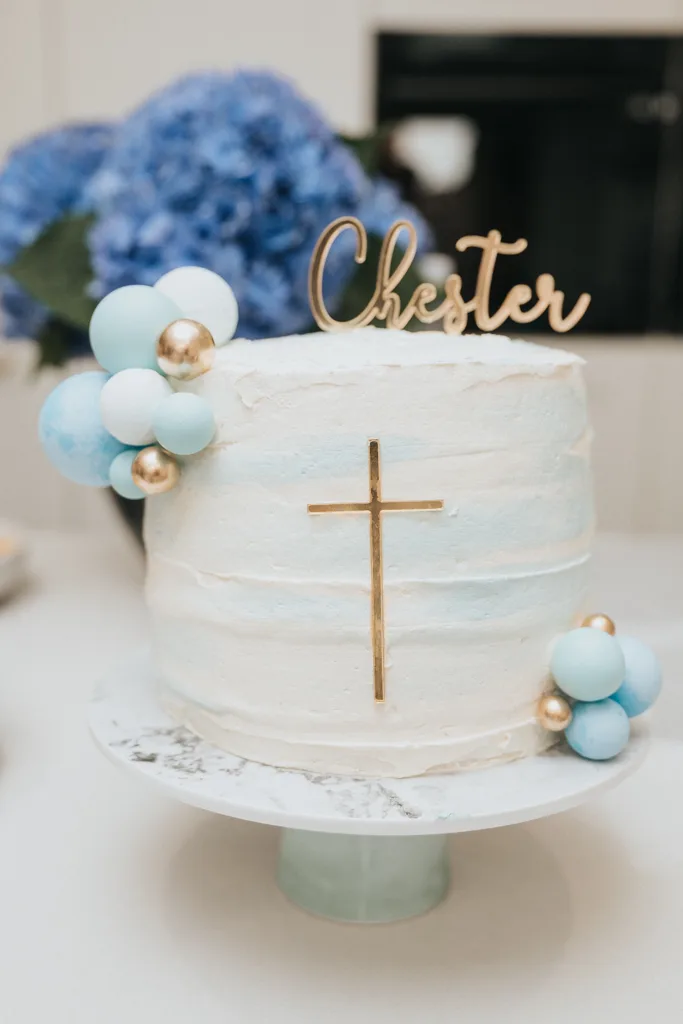 A round white cake with light blue accents, perfect for a christening in Waltham, features a gold cross on the side and a topper spelling "Chester." It is adorned with clusters of blue, white, and gold spheres. The cake sits on a white stand, with blurred blue hydrangeas in the background. © Aimee Lince Photography - Wedding photographer in Lincolnshire, Yorkshire & Nottinghamshire