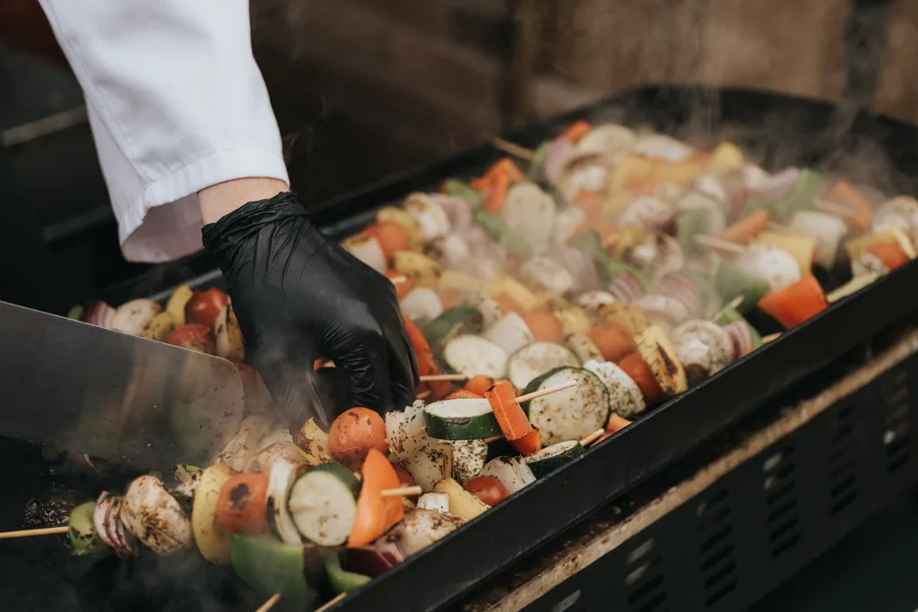 A person in a white coat and black glove expertly grills assorted vegetable skewers on a flat grill, with steam rising to signal the cooking progress. The vibrant mix includes mushrooms, zucchini, bell peppers, and onions—perhaps even prepared for a festive christening feast in Waltham. © Aimee Lince Photography - Wedding photographer in Lincolnshire, Yorkshire & Nottinghamshire