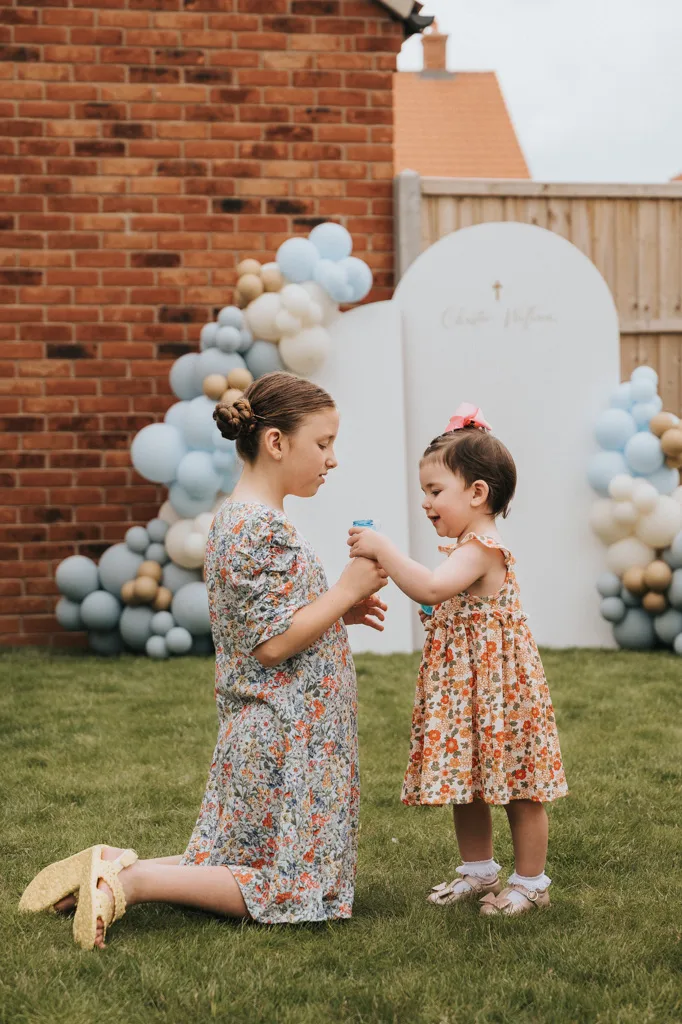 Two young girls in floral dresses play on the grass during a cheerful christening. The older girl kneels, holding a bottle, while the younger stands smiling with a pink bow. A backdrop of white and blue balloons rests against Grimsby's brick wall and wooden fence under a cloudy sky. © Aimee Lince Photography - Wedding photographer in Lincolnshire, Yorkshire & Nottinghamshire